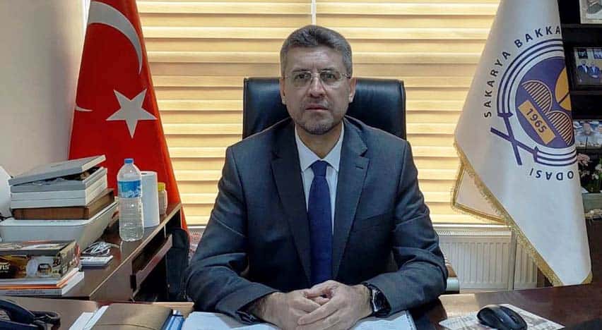 Yönetici ofisinde oturan bir erkek, arka planda Türk bayrağı ve basın merkezi bayrağı ile resmi orta.